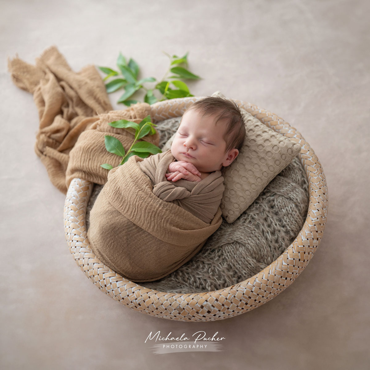 Schlafendes Neugeborenes, in warme beigefarbene Tücher gewickelt, liegt in einem runden Korb auf weichen Stoffen mit kleinen grünen Blättern als Akzent.